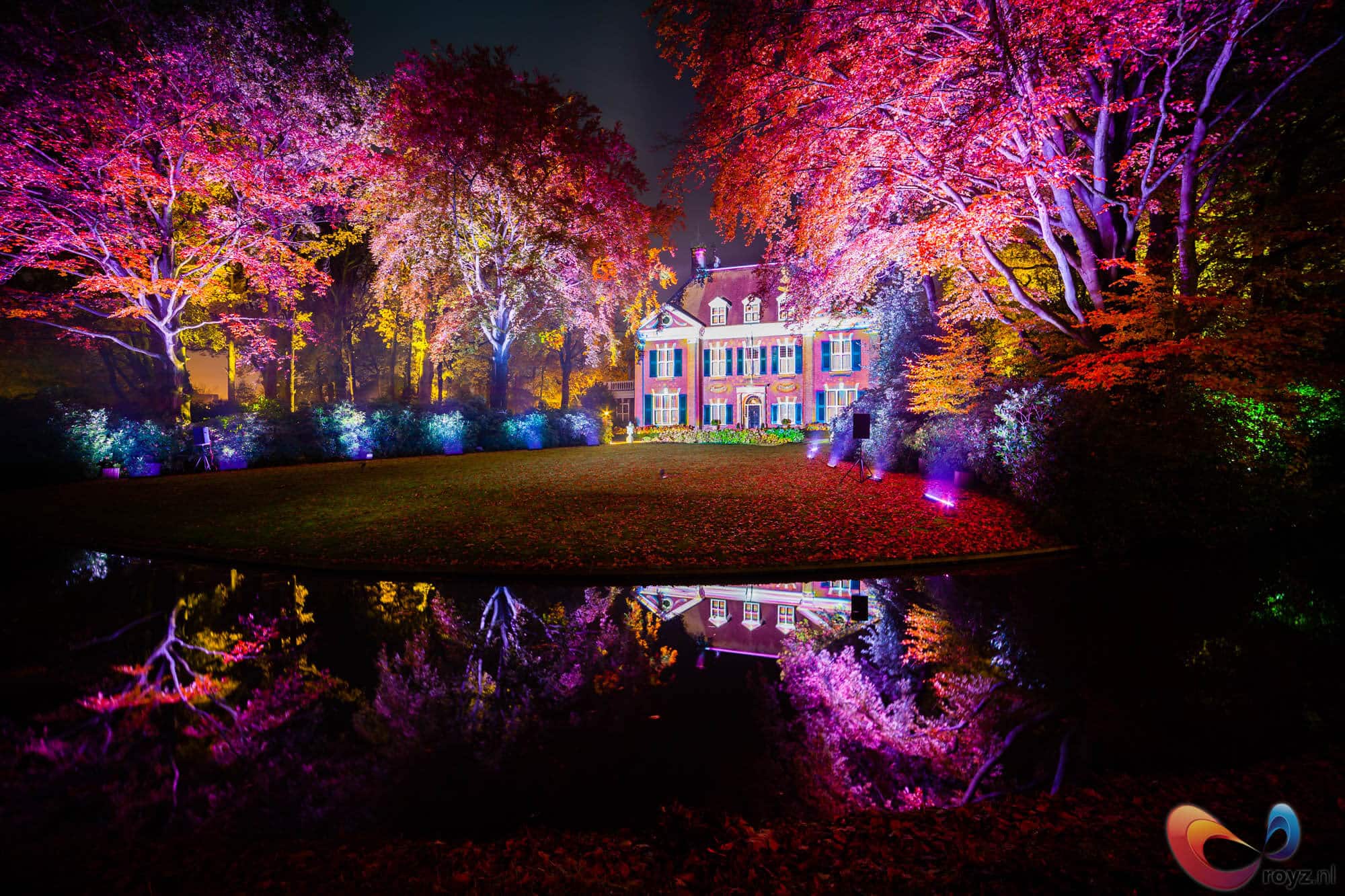 Neonverlicht landhuis met waterreflectie en kleurrijke herfstbomen in nachtelijk tuinlandschap.