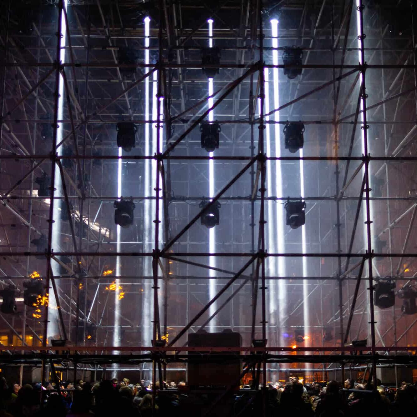 Grote stalen steigers met neonlichten, publiek als silhouetten bij nachtconcerten.