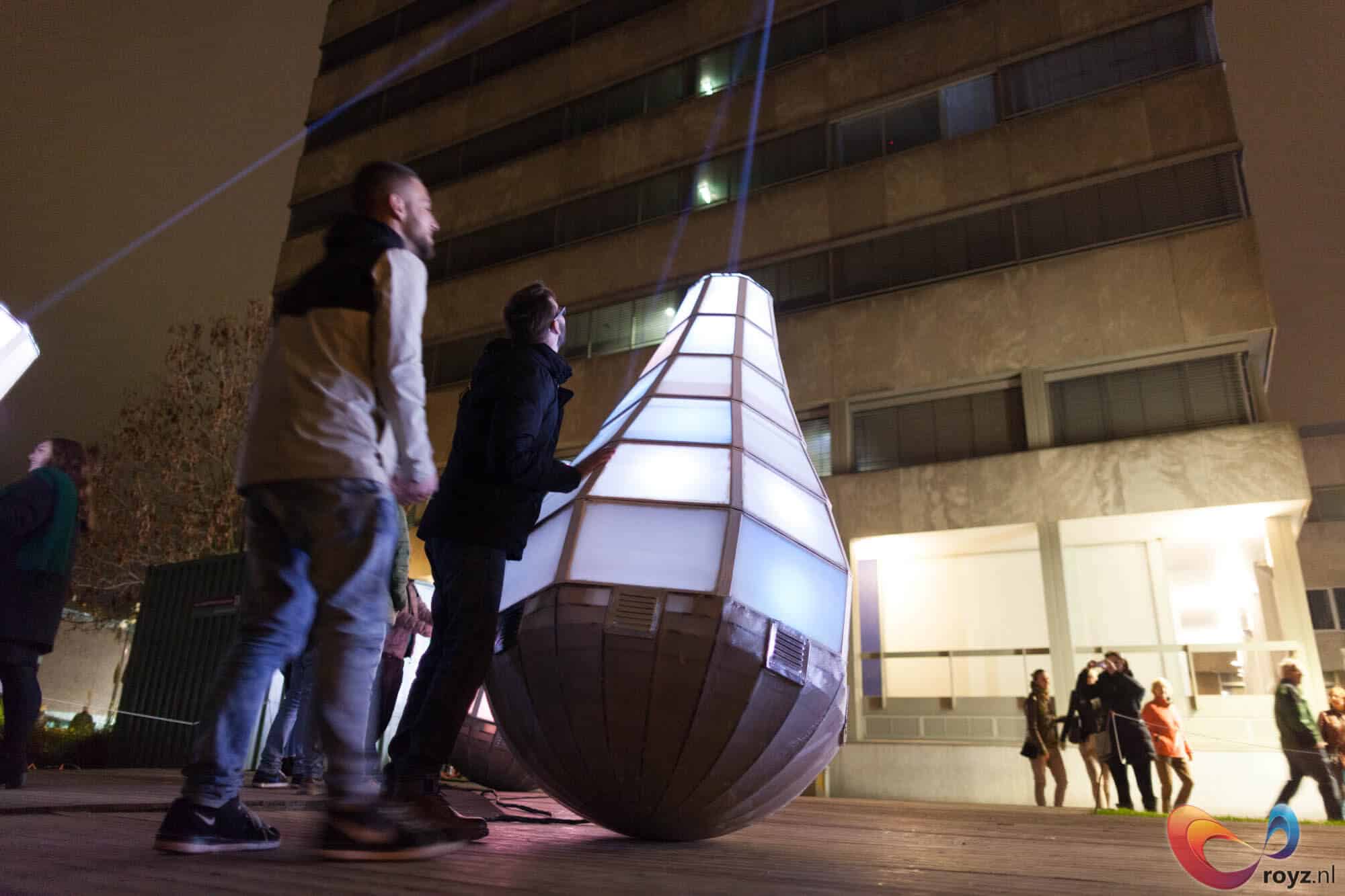 Futuristische teardrop-sculptuur verlicht avondplein Eindhoven met gloeiend bovenwerk en ribbelige onderkant; omstanders kijken.