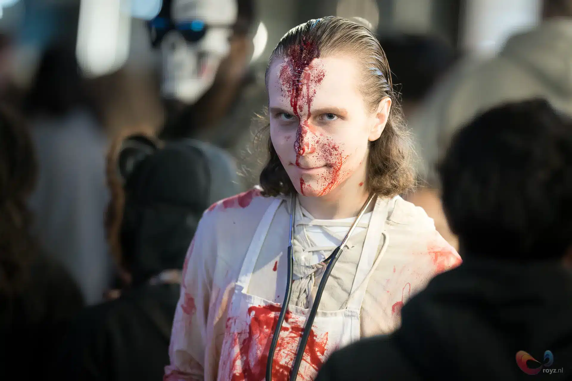 Horror cosplay met bloedstreep op gezicht en witte schort, koude blik, publiek op achtergrond.