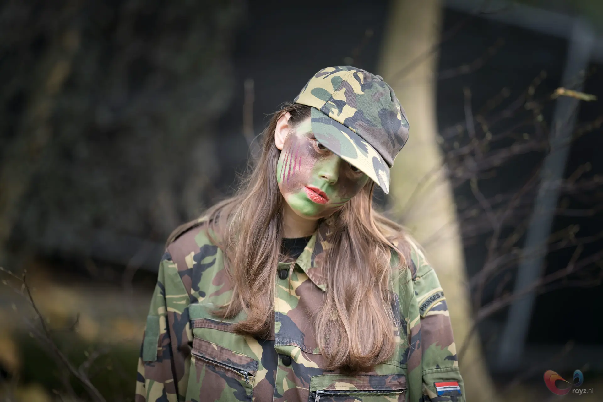 Vrouw in camouflagekleding met groene gezichtsschmink en kleurrijke lijnen op wangen, rode lippen in bos.
