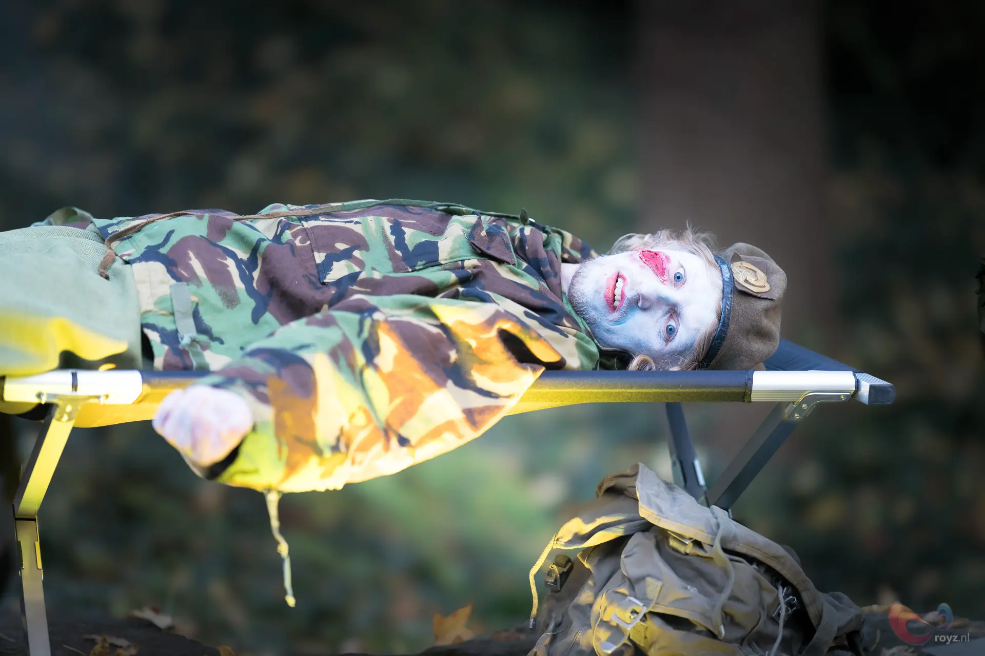 Spookachtige soldaat in camouflage op veldbed in bos, blauw gezicht en rode lippen.