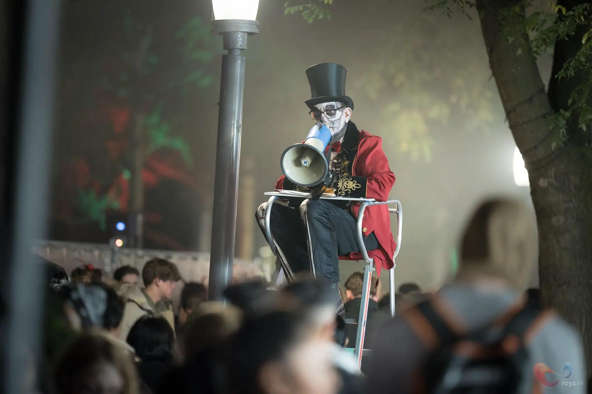 Straatartiest in rode tailcoat op hoge witte stoel, wit geschminkt, met blauwe megafoon tijdens stadsavond.