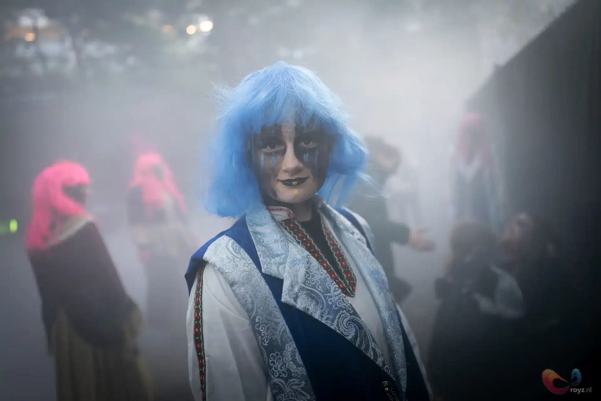 Performer met felblauwe bob-wig, porseleinen huid en damastvest met zilveren patronen op maskerbal.