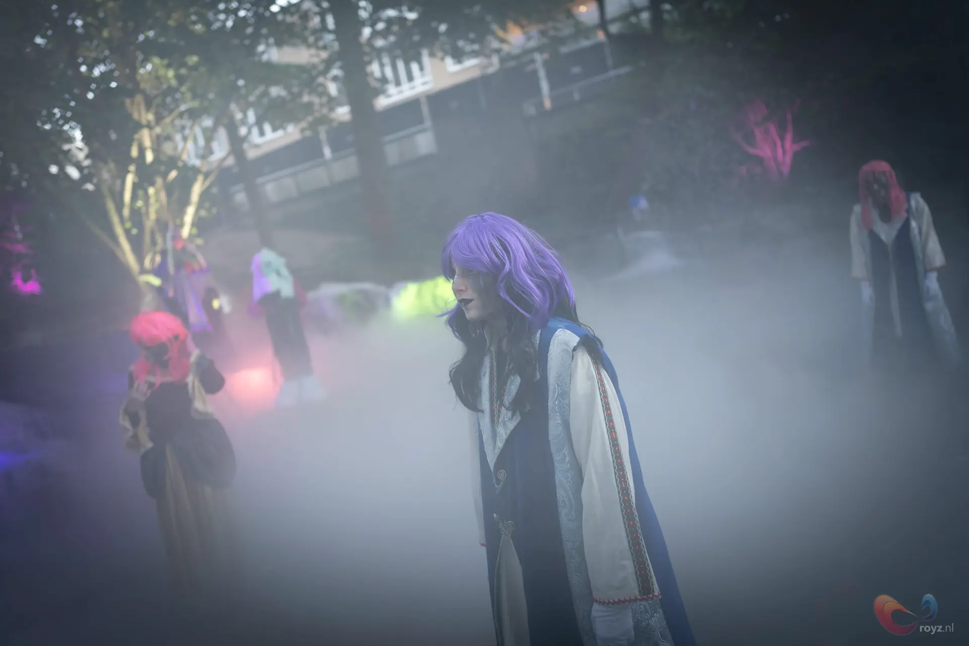 Centraal figuur met blauwe cape en paarse pruik in nevelige cosplay-scène met neonlichten.