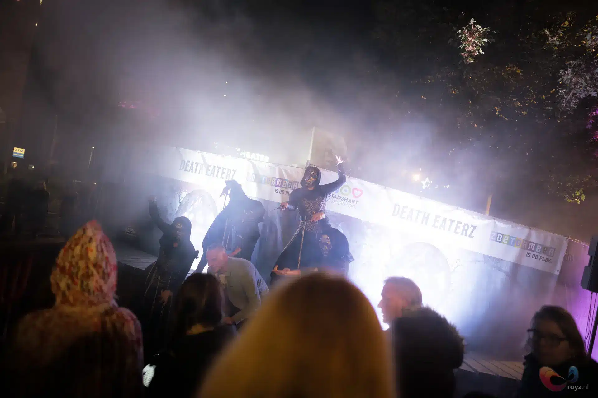 Nachtelijk silhouet op podium met bord omhoog, rook en lange witte banner, publiek kijkt gespannen.