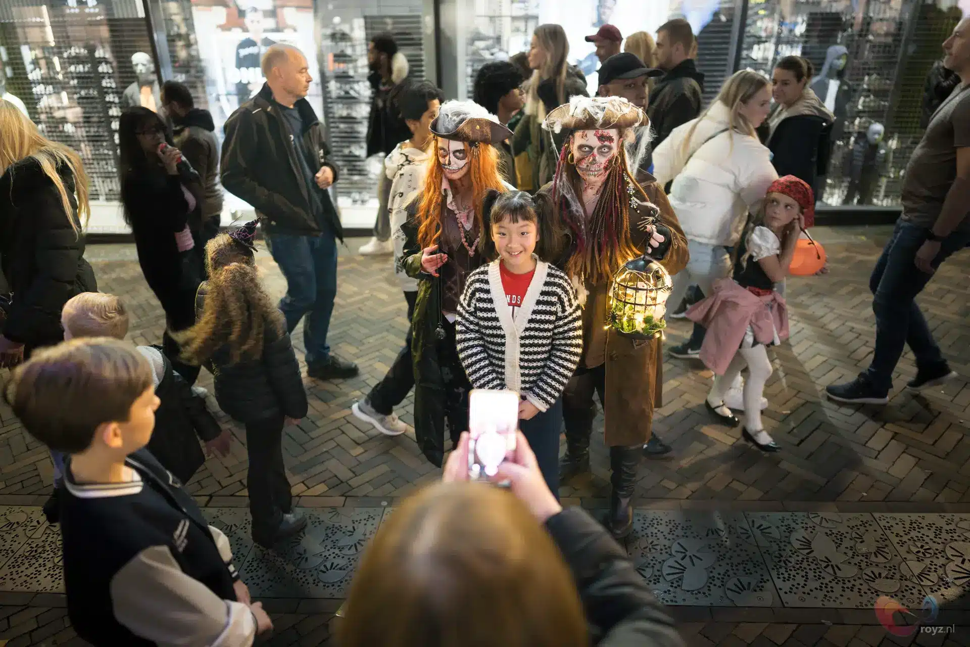 Kind poseert tussen twee straatartiesten met lantaarn op drukke Halloween winkelstraat.