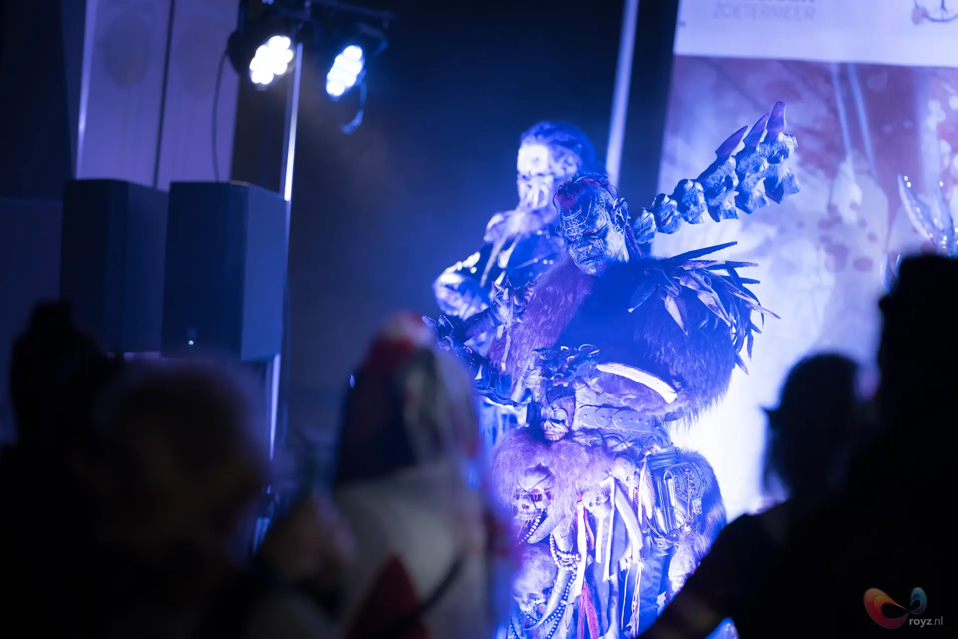 Monsterkostuum met botpantser en schedelmotieven op blauw podium, artiest met bleke schedelmake-up en rook.