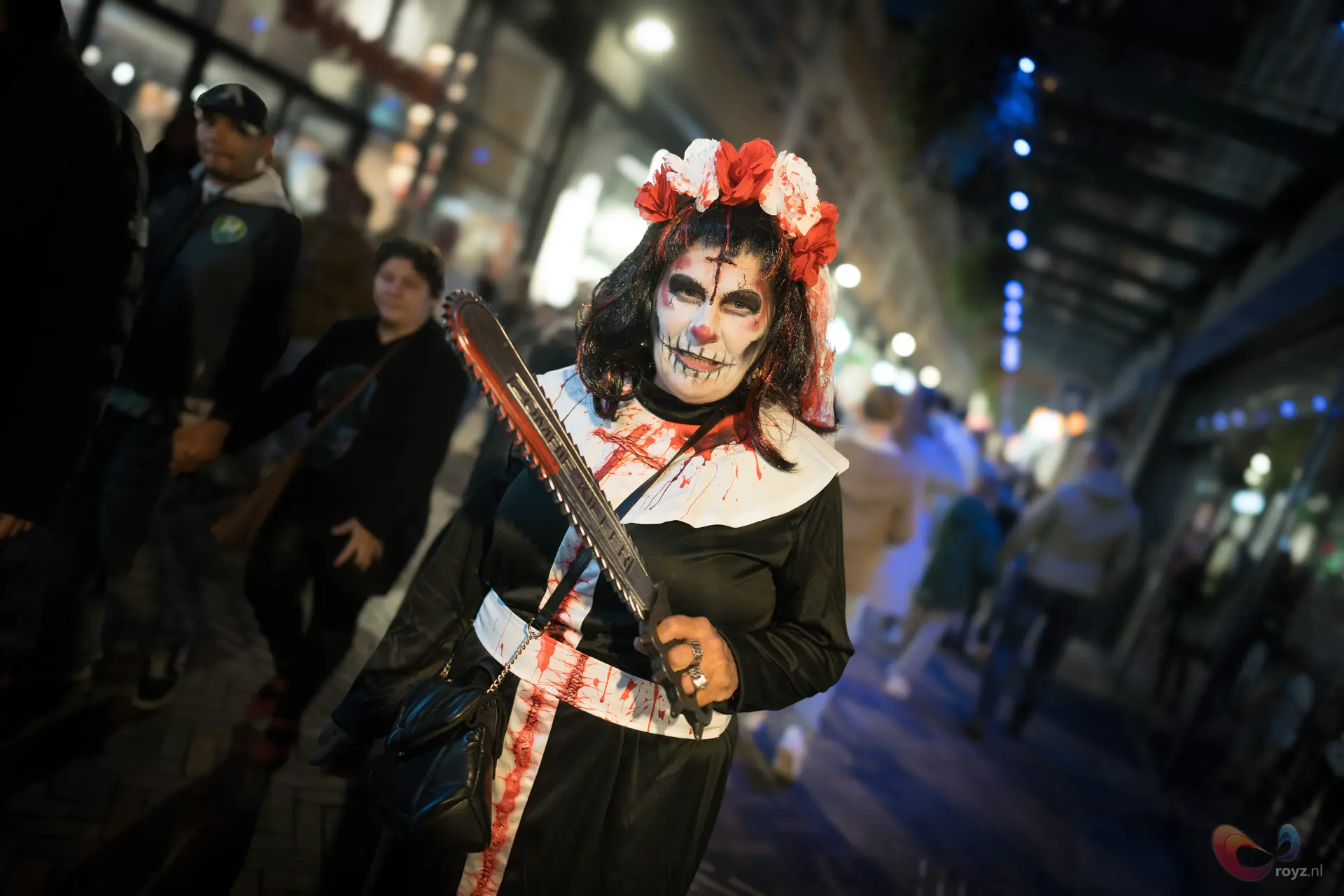 Zoetermeer Halloween figuur met skeletmake-up, lange zwarte jurk, bloemkroon, kettingzaag bij neonlichten.