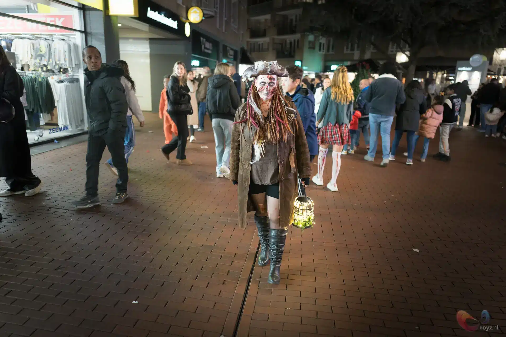 Kostuumfiguur met groene lantaarn in drukke avondstraat vol publiek.