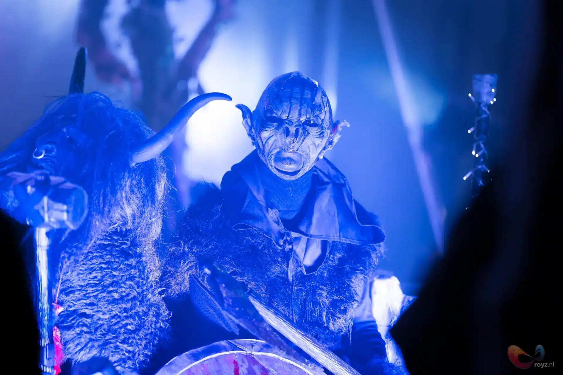 Demonmasker in blauw theaterlicht; harige mantel en ritueel mythisch tafereel, hoornfiguur nadert met staf.