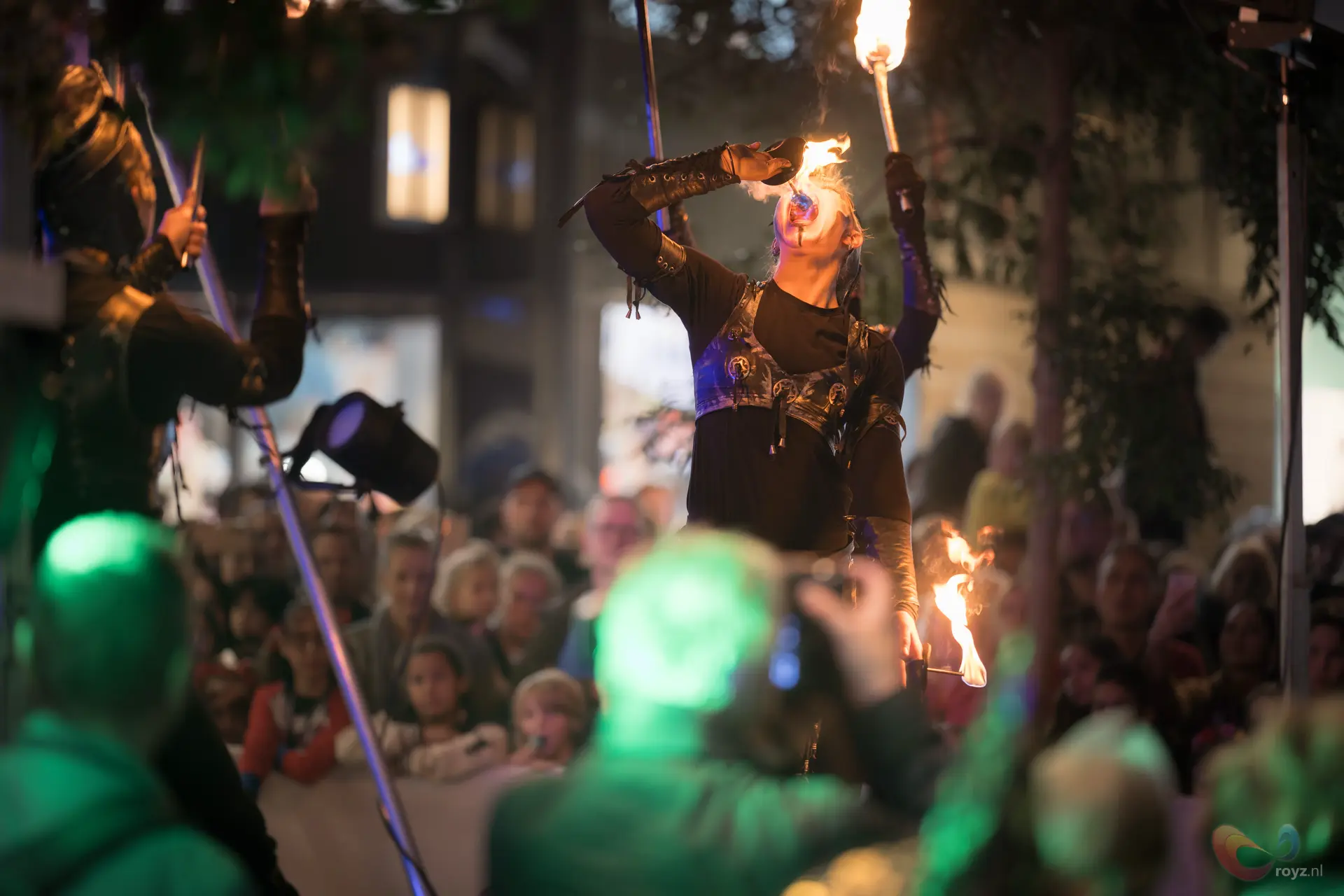 Nachtelijke vuurperformer op straat met mondvlam en brandende stokken, publiek ademloos.