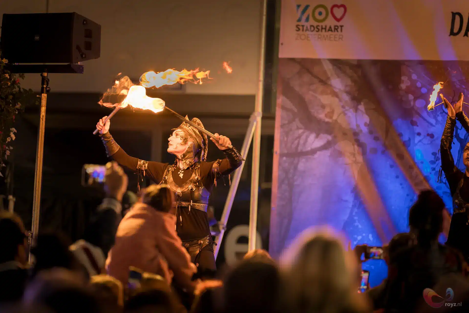 Vrouwelijke vuurperformer danst met twee fakkels op nachtelijk festivalpodium, publiek als silhouetten.