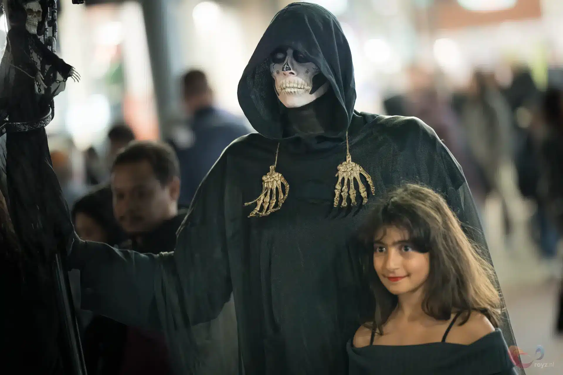 Cosplayduo: grimmige doodsfiguur met schedelmasker en gouden skeletachtige hangers naast glimlachend meisje.