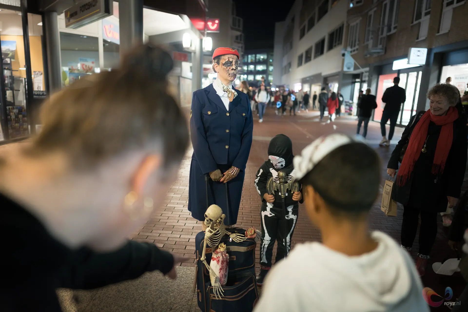Levende standbeeld skeletmake-up in donkerblauw uniform, rode pet, skeletprops en kind bij Zoetermeer nachtmarkt.