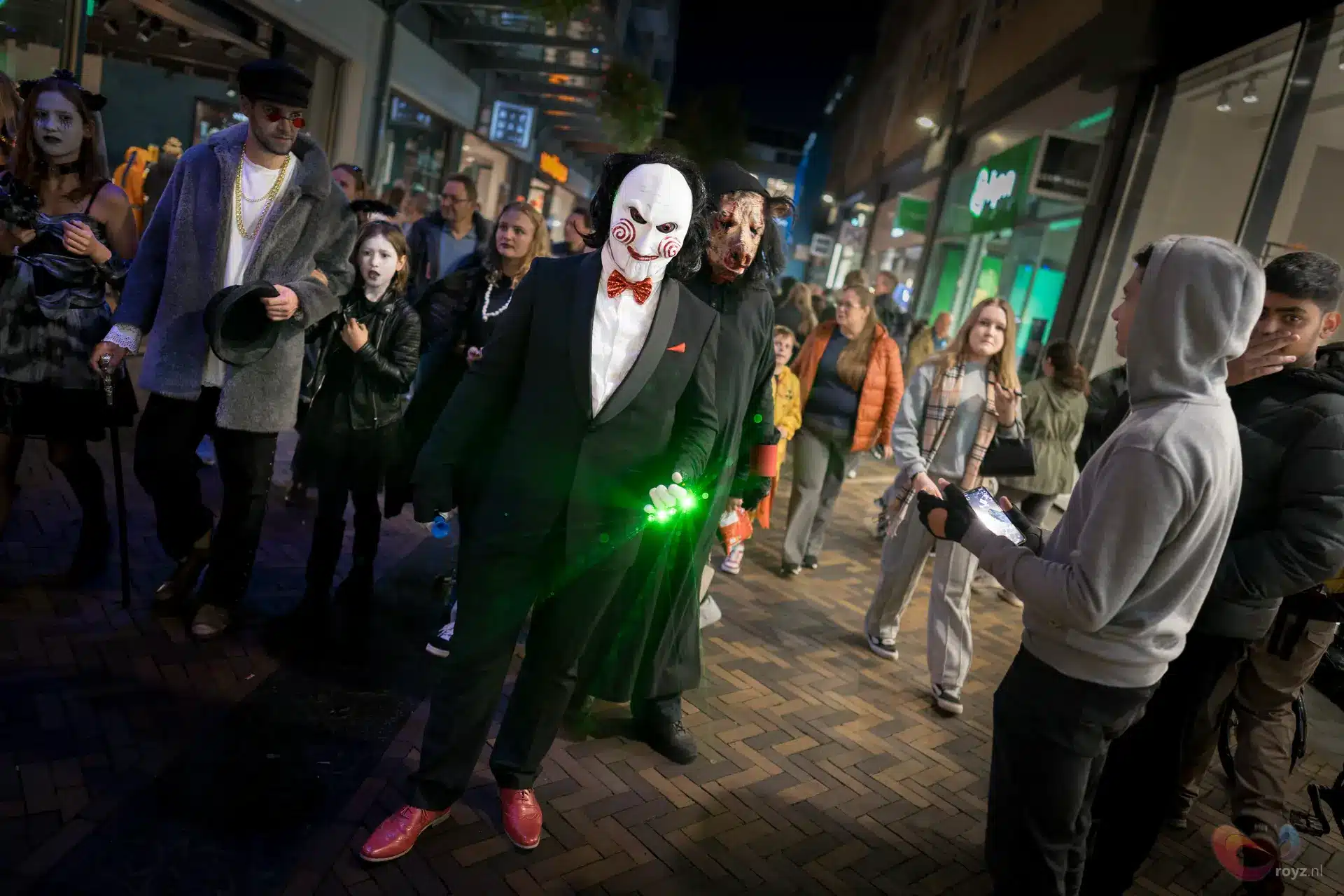 Maskerduo Halloween Zoetermeer: elegante man met groene gloeiende prop naast groteske partner en nieuwsgierige toeschouwers