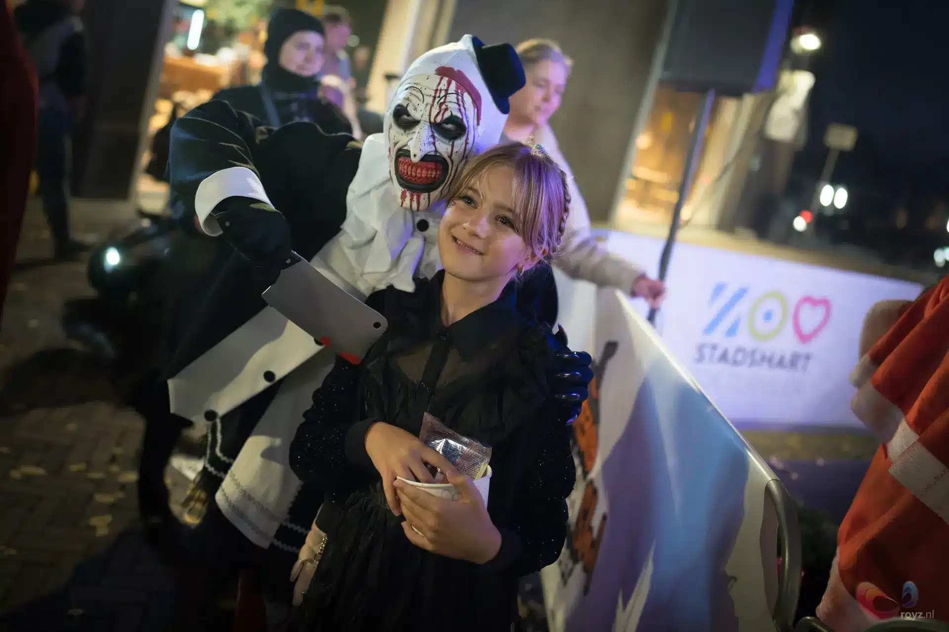 Halloween in Zoetermeer met gemaskerde figuur en meisje onder warme straatverlichting.