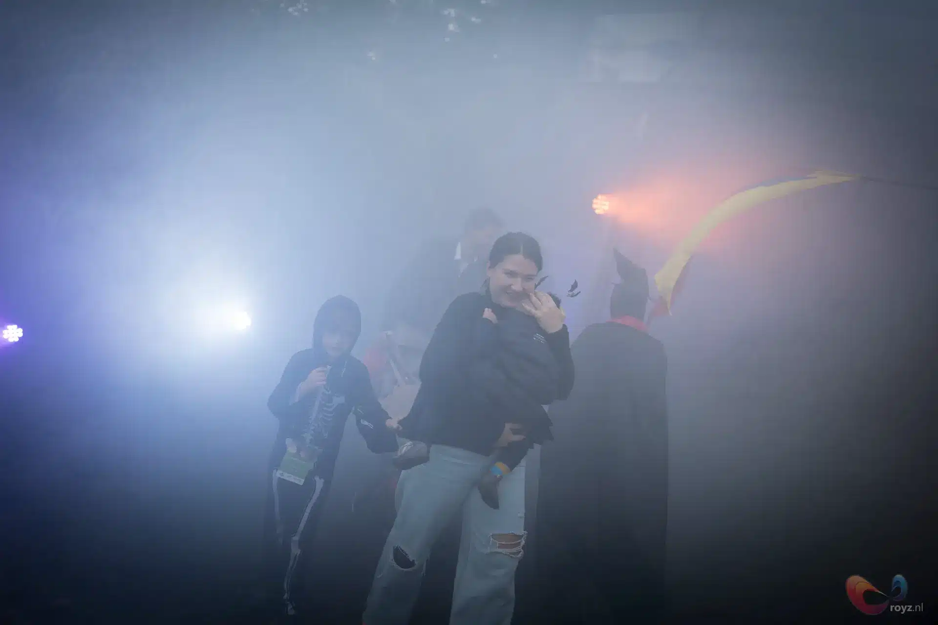 Nevelig podium tijdens optreden met artiesten: man in hoodie, gitarist en mantelfiguur met oranje gloed
