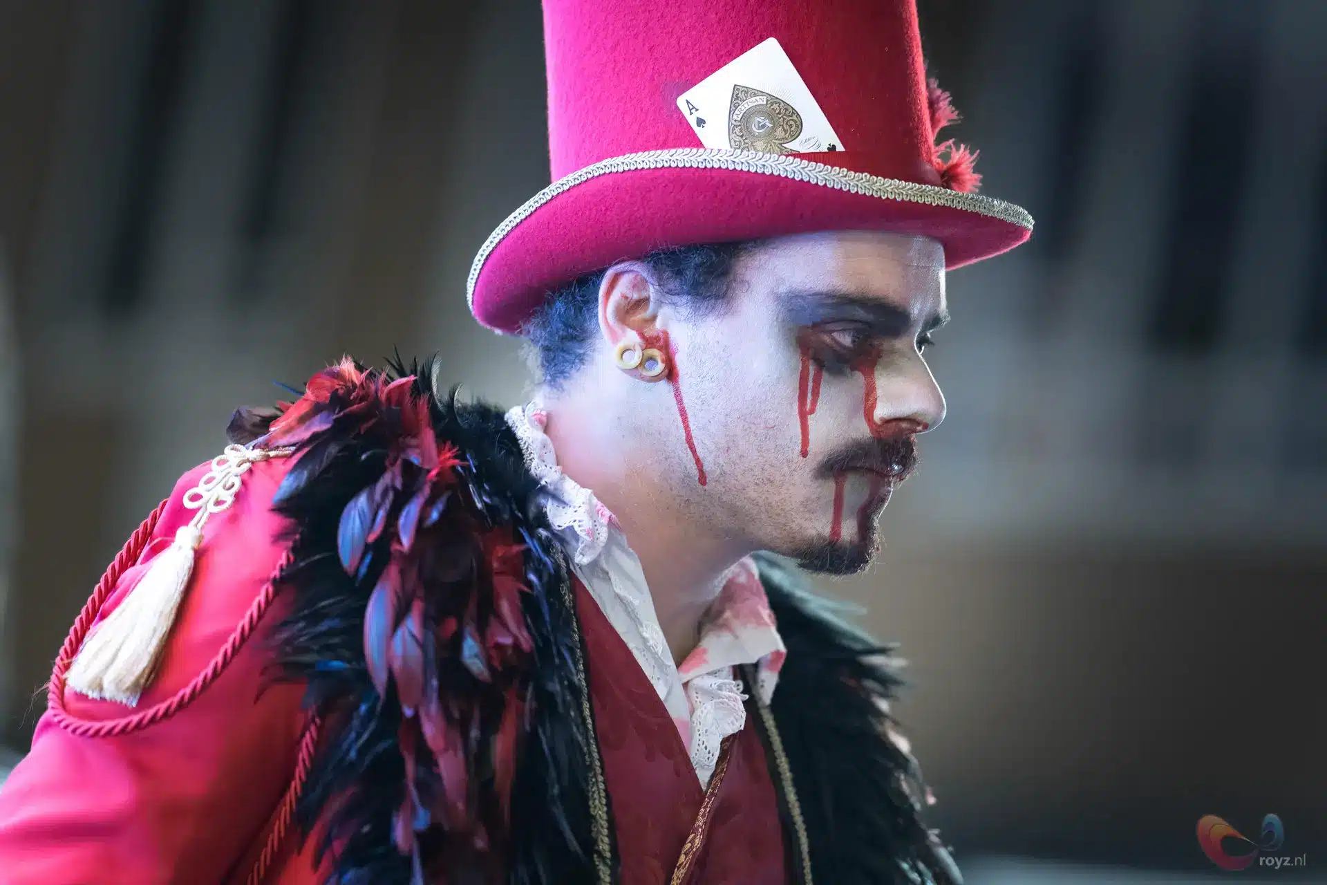 Pierrotachtige performer in carnavalsnacht met felroze hoed, rood jasje, gouden koorden en melancholische make-up.