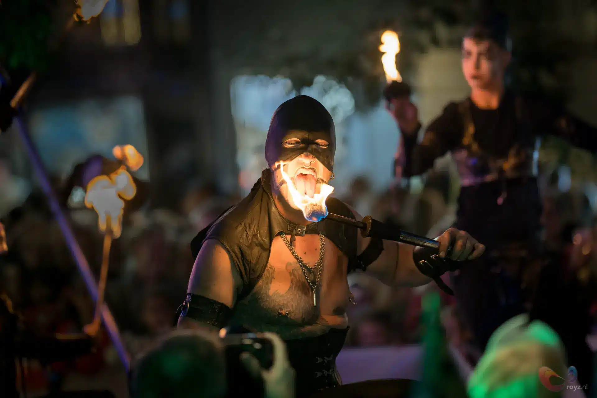 Vuurspuwer met masker spuwt vlam op nachtfestival; silhouetten en fakkels.