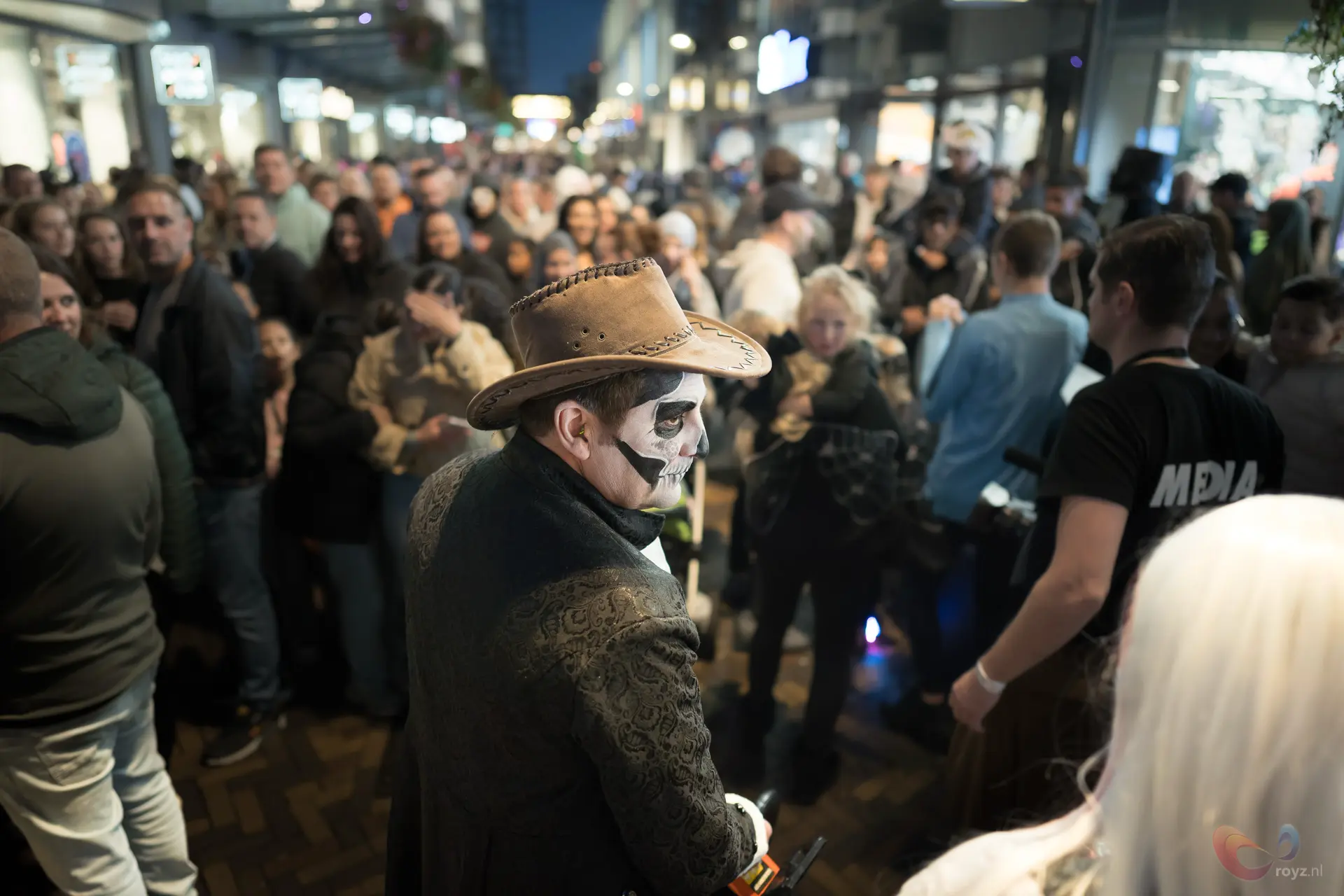 Skeletmime met cowboyhoed in druk winkelcentrum; media filmen terwijl publiek sfeer vastlegt.