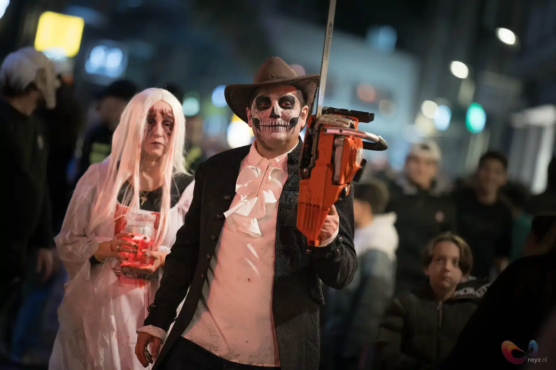 Zoetermeer Halloween: schedelgezicht met oranje kettingzaag, spookvrouw in roze jurk, neonverlichting en straatparade.