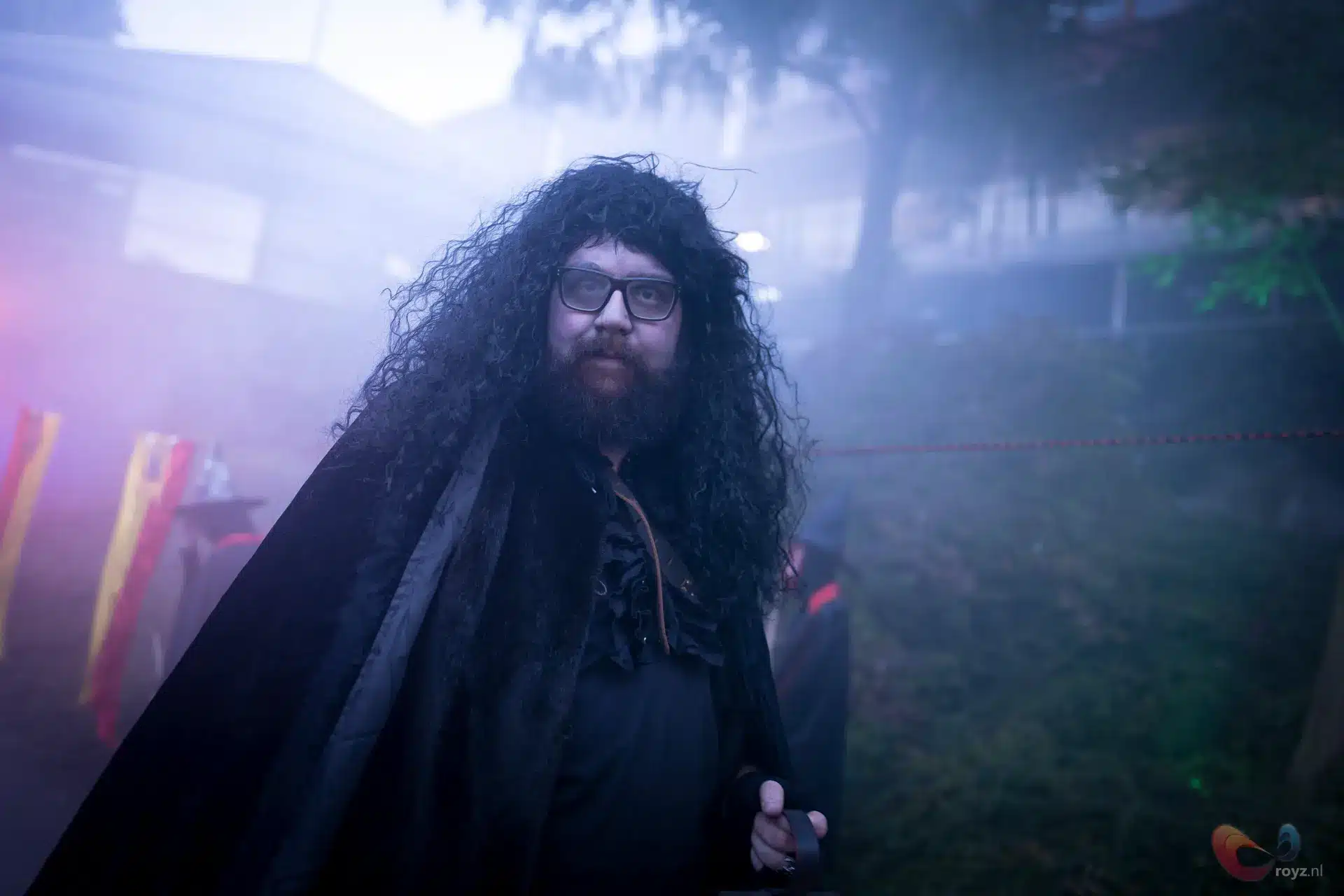 Tovenaar met lange krullende haren en baard in schemerlicht, lavendelnevel en rode gele linten.