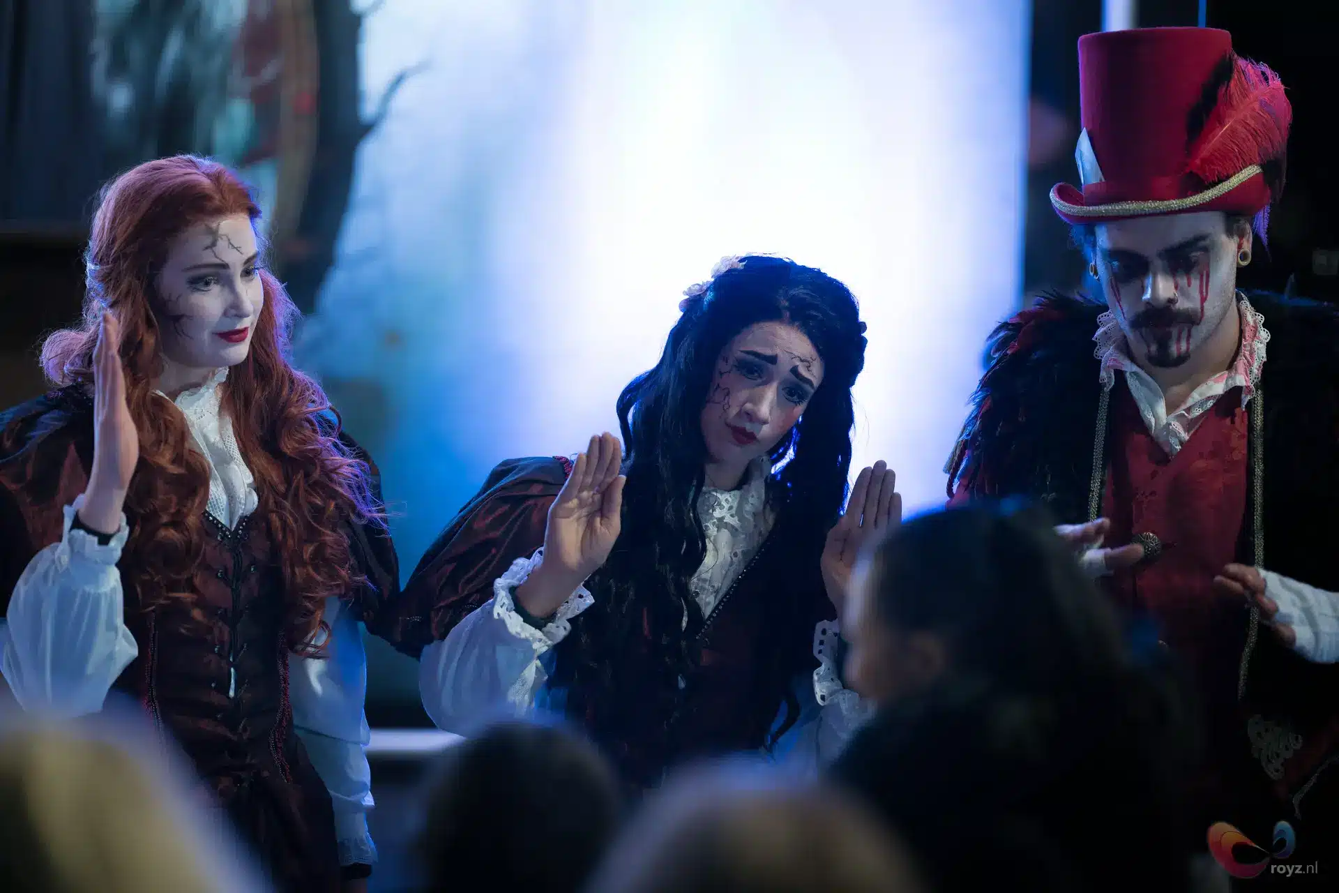 Drie gothische figuren in donker theater met koel blauw licht en carnavalachtige sfeer.