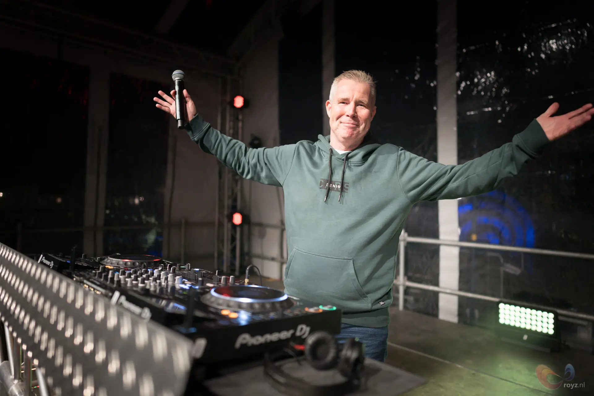 DJ-host op podium met mixer en platines onder rode clubverlichting.