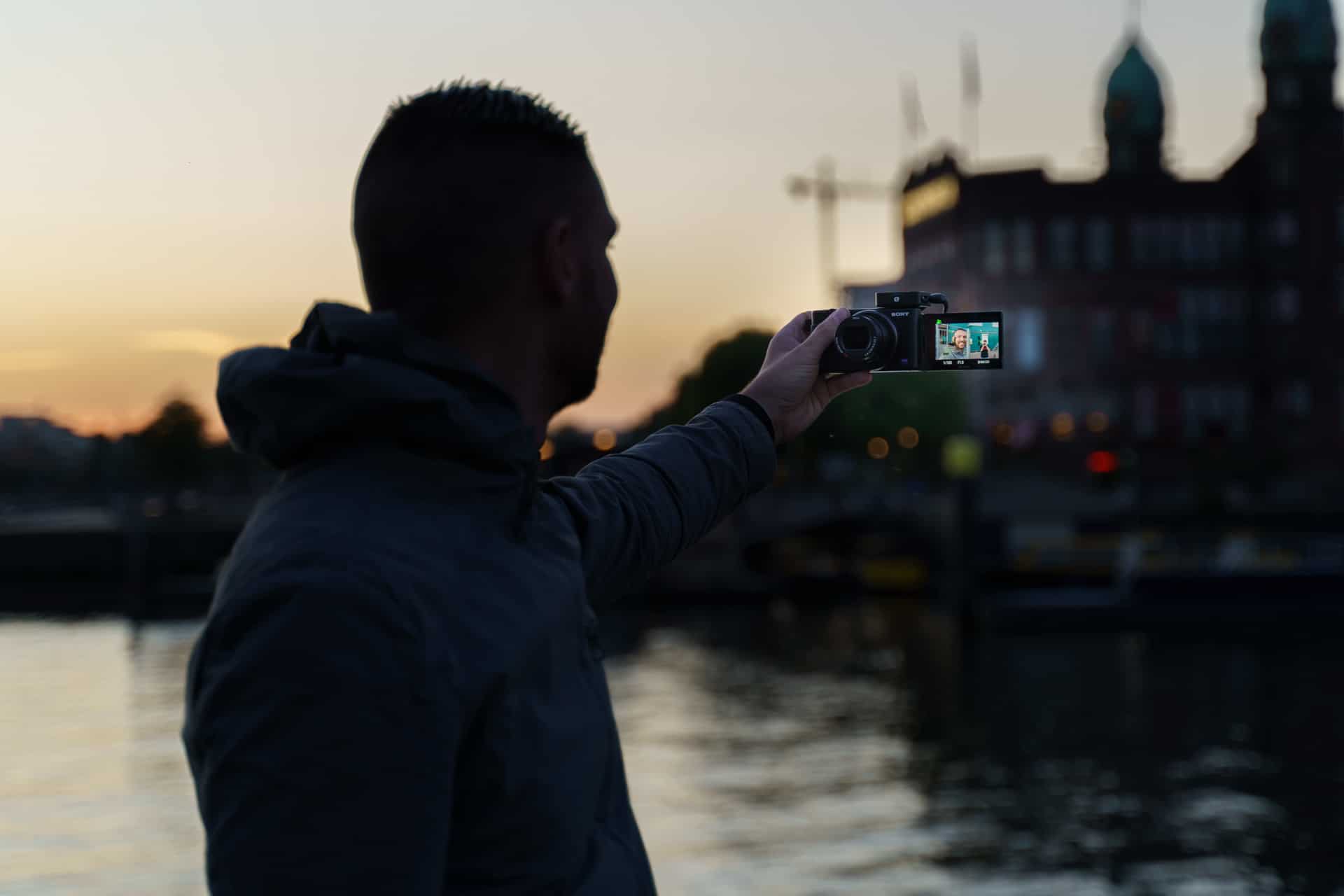 Fotograaf silhouet bij zonsondergang aan rivier met compacte Sony camera en stadszicht