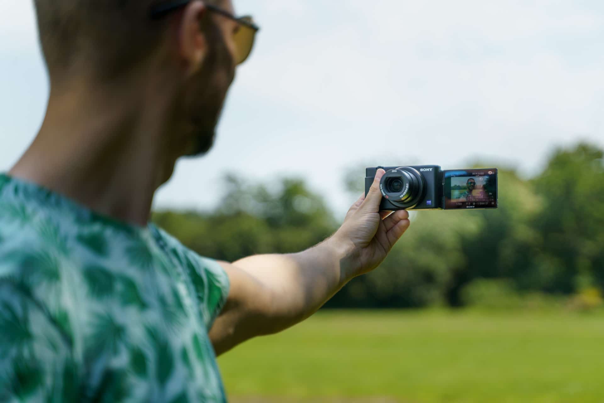 Buiten vloggen met compacte Sony-camera, groene palmbladpatroon shirt, zonnig veld.