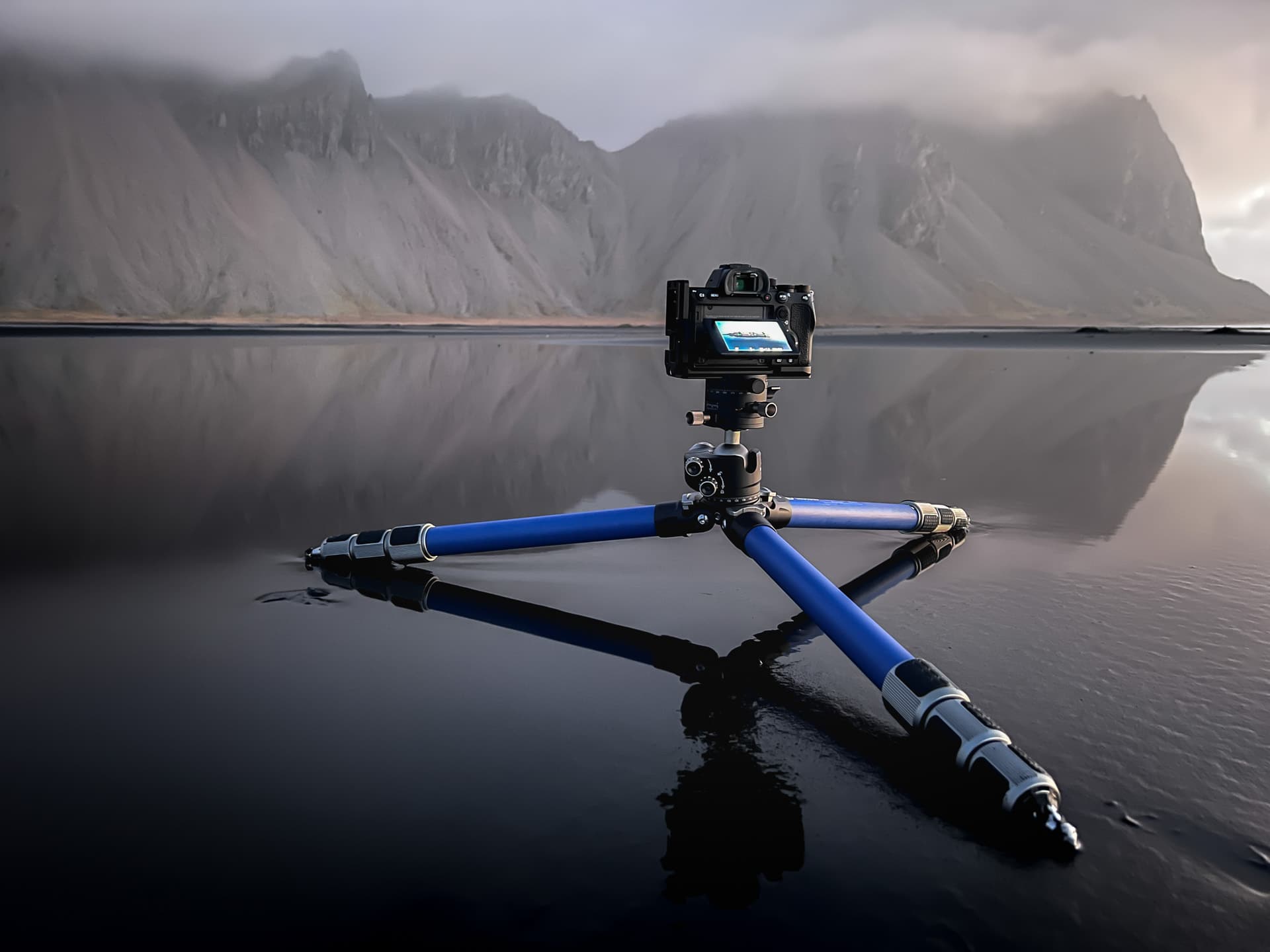 Compacte camera op blauw statief boven spiegelend water met berglandschap en ochtendlicht.