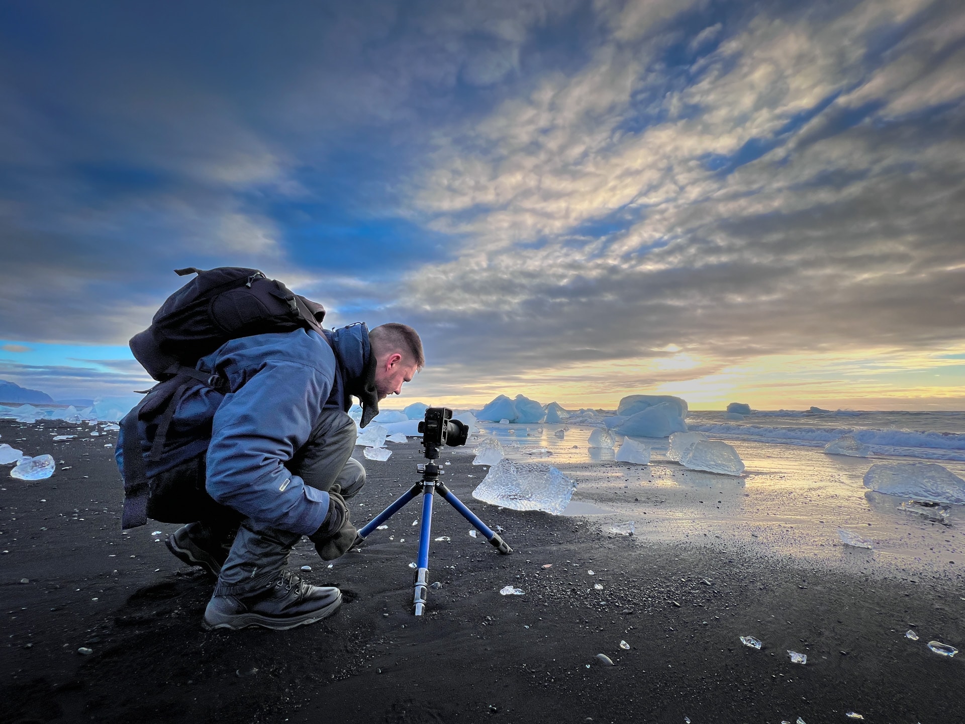Fotograaf met statief en camera op zwart zandstrand bij Jökulsárlón, ijsblokken en zonsondergang.