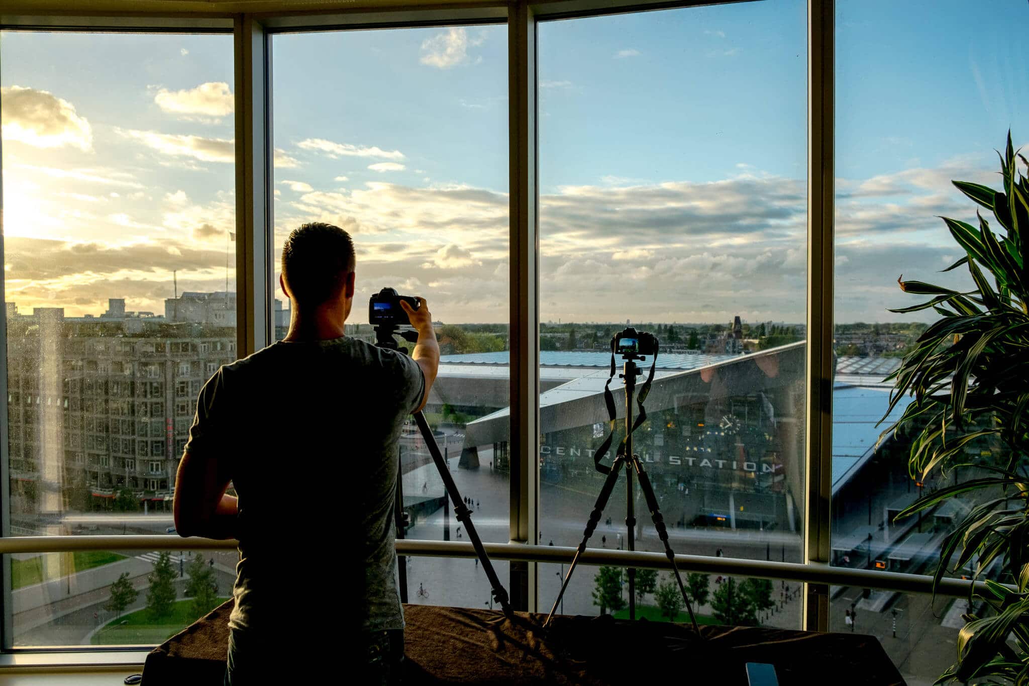 Fotograaf in moderne studio met grote ramen, stadszicht en twee tripods bij zonsondergang.