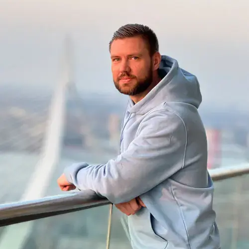Man met lichtblauwe hoodie bij glazen railing, brug en rivier in gouden licht.