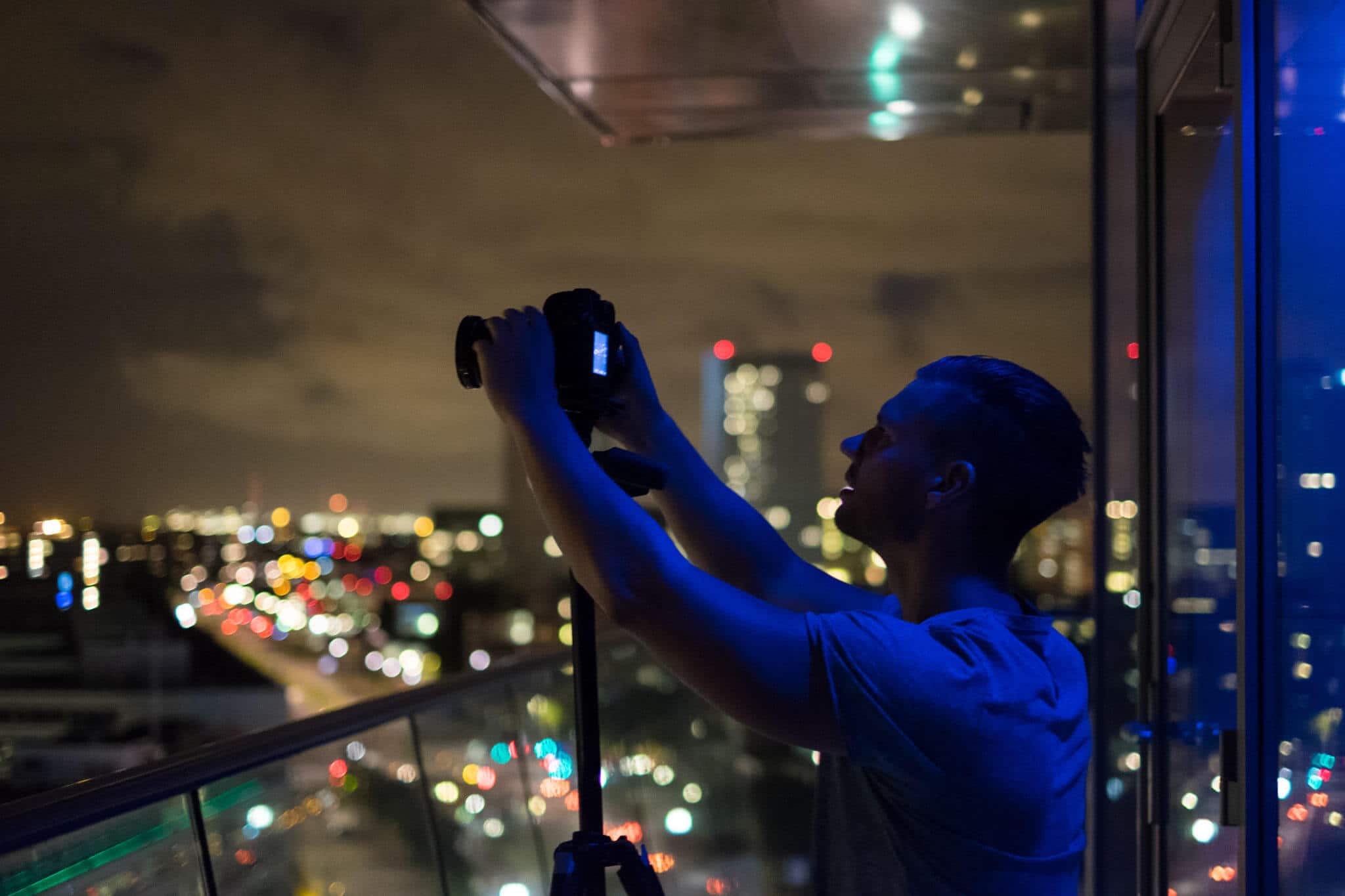 Fotograaf in blauw T-shirt bereidt camera op balkon met glazen balustrade boven nachtelijke stadslichten.