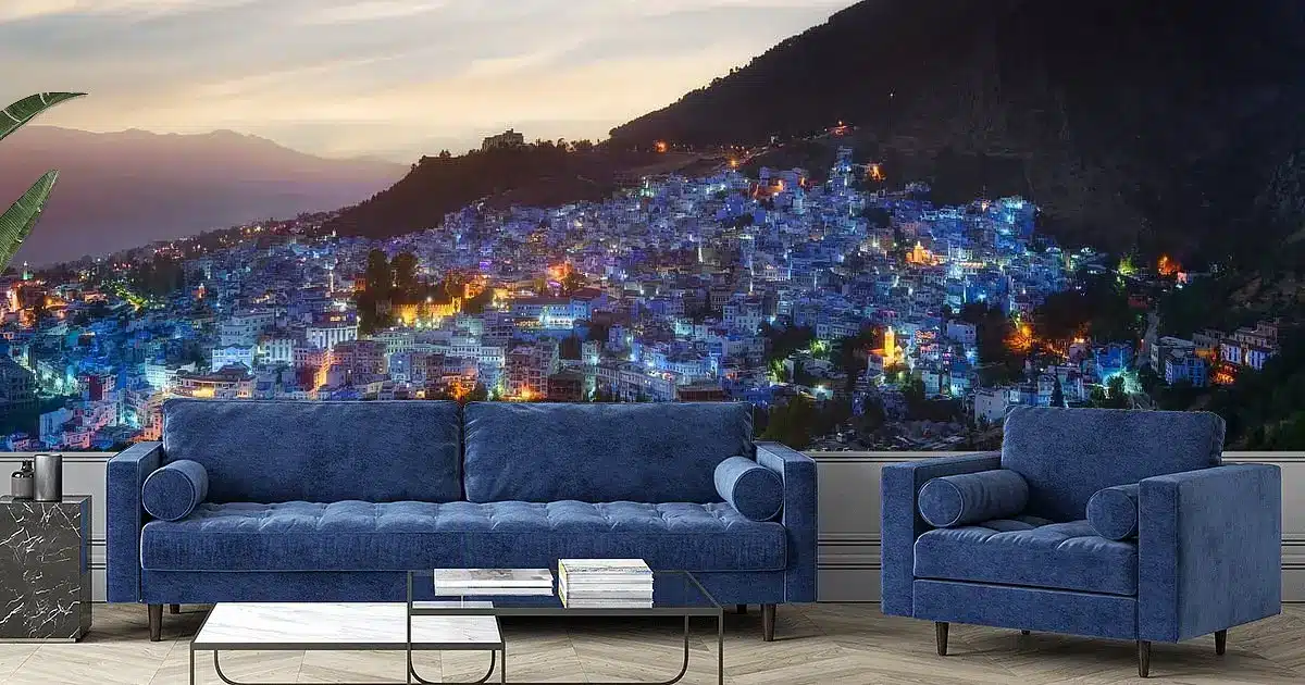 Chefchaouen blauw muurschilderij in woonkamer met velvet bank en fauteuil bij schemering en glazen salontafel.