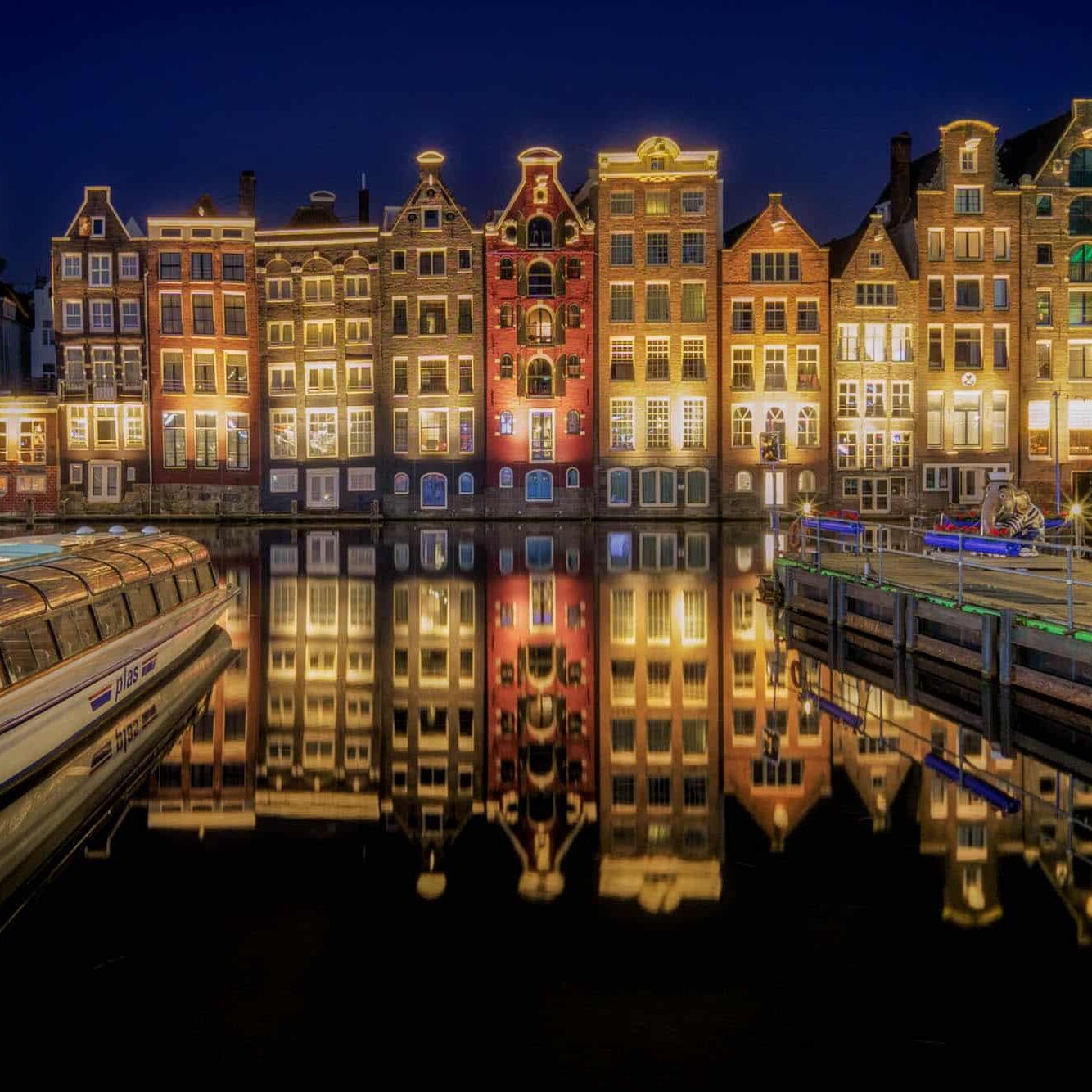 Nacht langs Amsterdamse grachten met glazen dekboot, warme ramen en neonlichten, reflecties in kalm water.