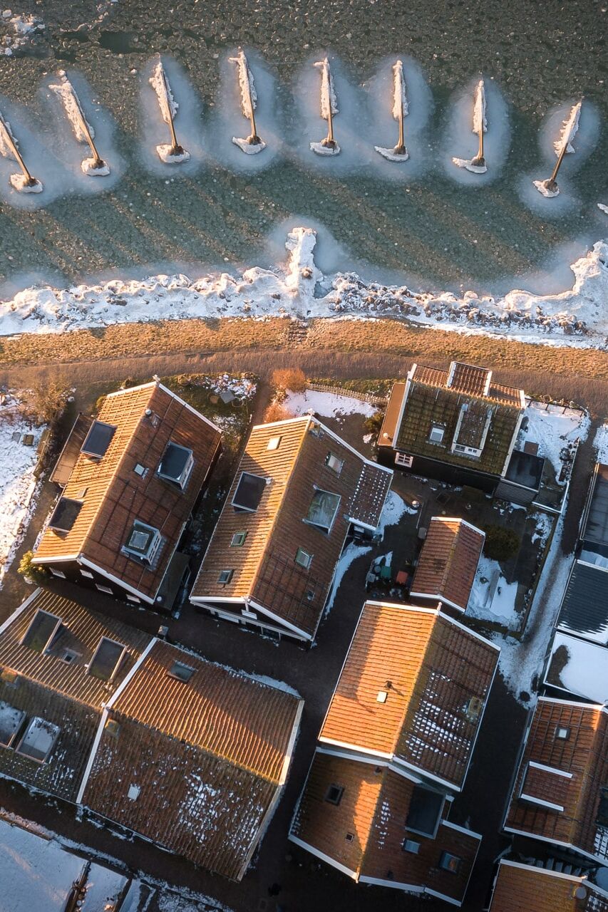 Luchtfoto van een winterse kustwijk met oranje daken, sneeuw en golfbrekerpalen langs ijswater.