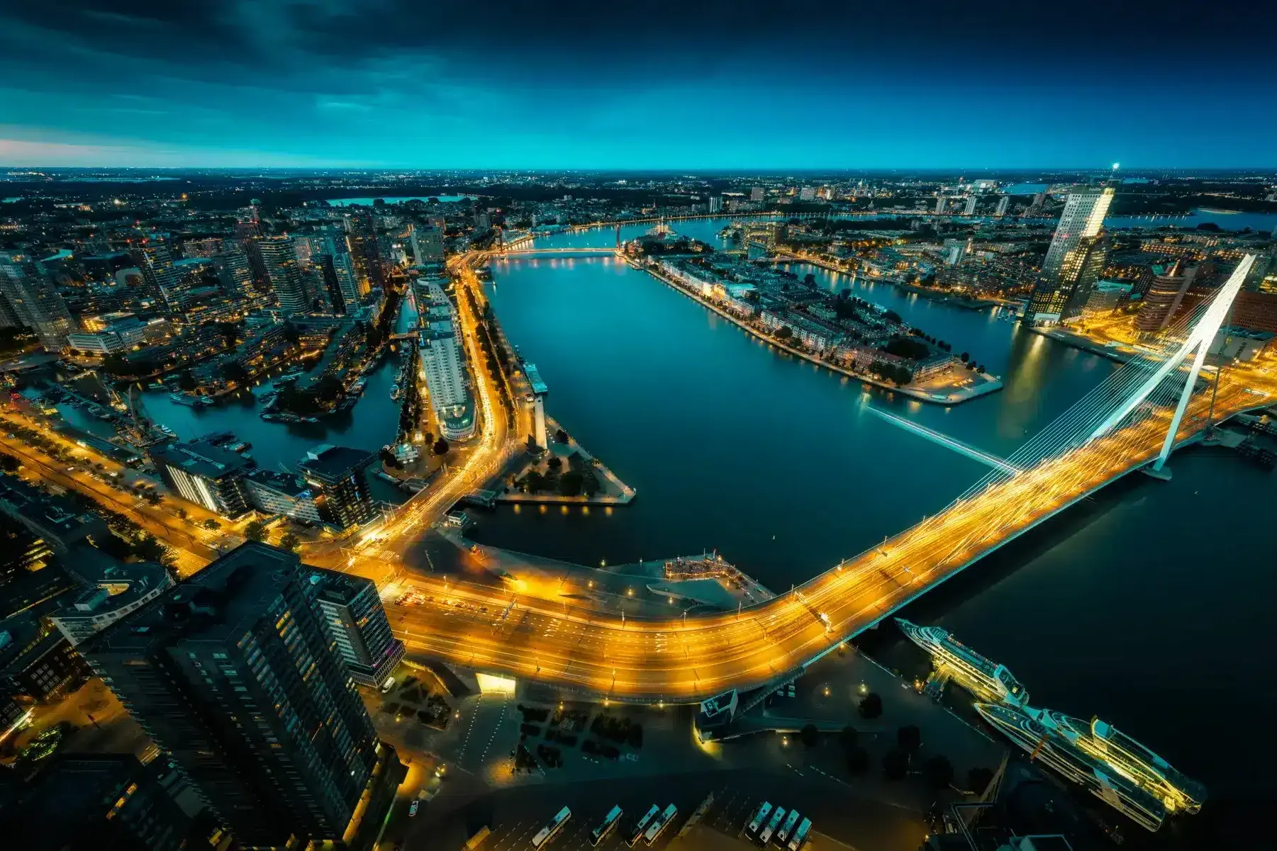 Nachtzicht op een witte kabelbrug boven een havenstad met amberkleurige verlichting en waterreflecties.
