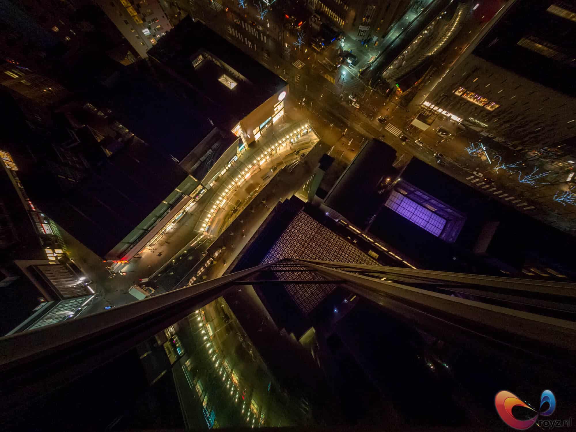 Nachtelijke skyline met amberkleurige straatverlichting, reflecties op natte straten, glazen railing en paarsachtig blauw licht.
