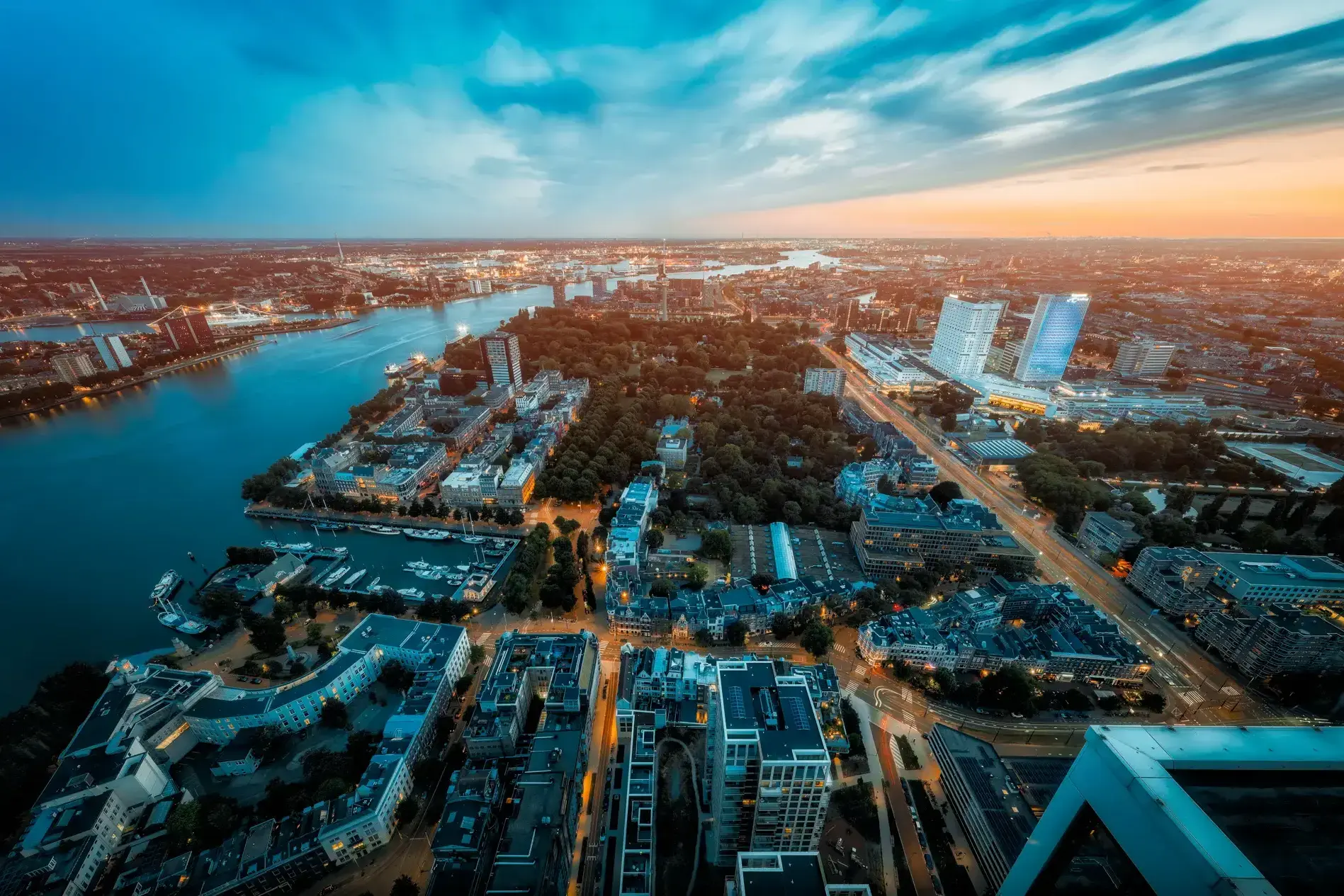 Rivierfront met groen park, marina en hoogbouw langs water bij zonsondergang.