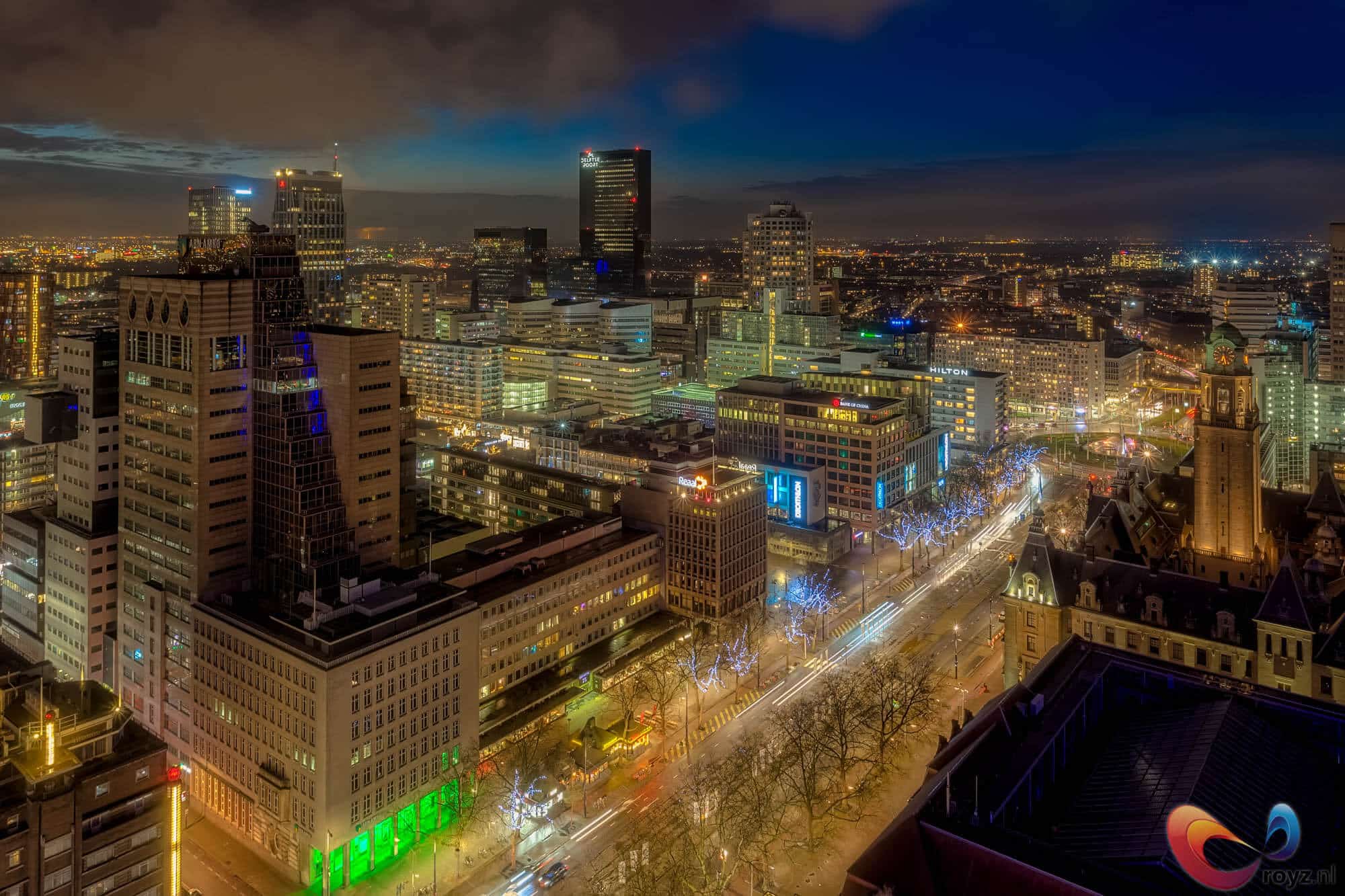 Nachtelijke skyline met Hilton-sign en kloktoren, lange verkeersbelichting, blauwe kerstverlichting langs bomen.