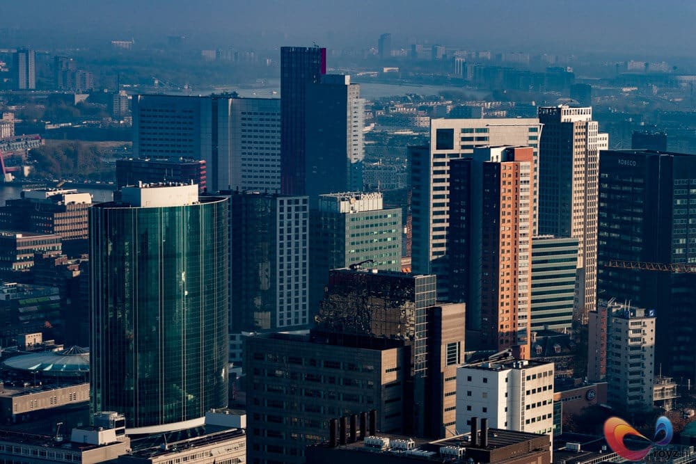 Dichte stedelijke skyline met cilindrische groene glazen toren, nevel en zonreflecties op glas en beton.