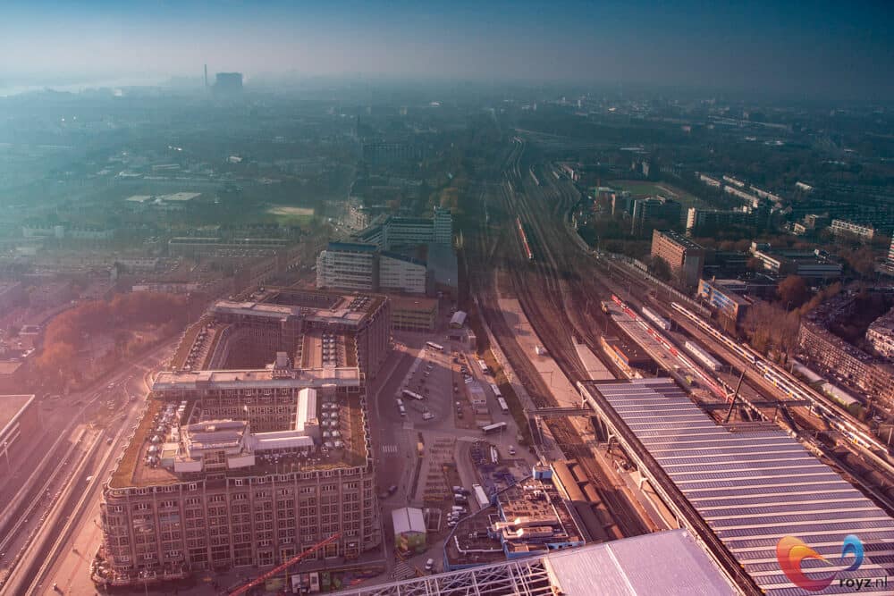 Dageraad op een stedelijk spoorwegknooppunt met overkappingen, treinen langs perrons en blokvormige gebouwen.