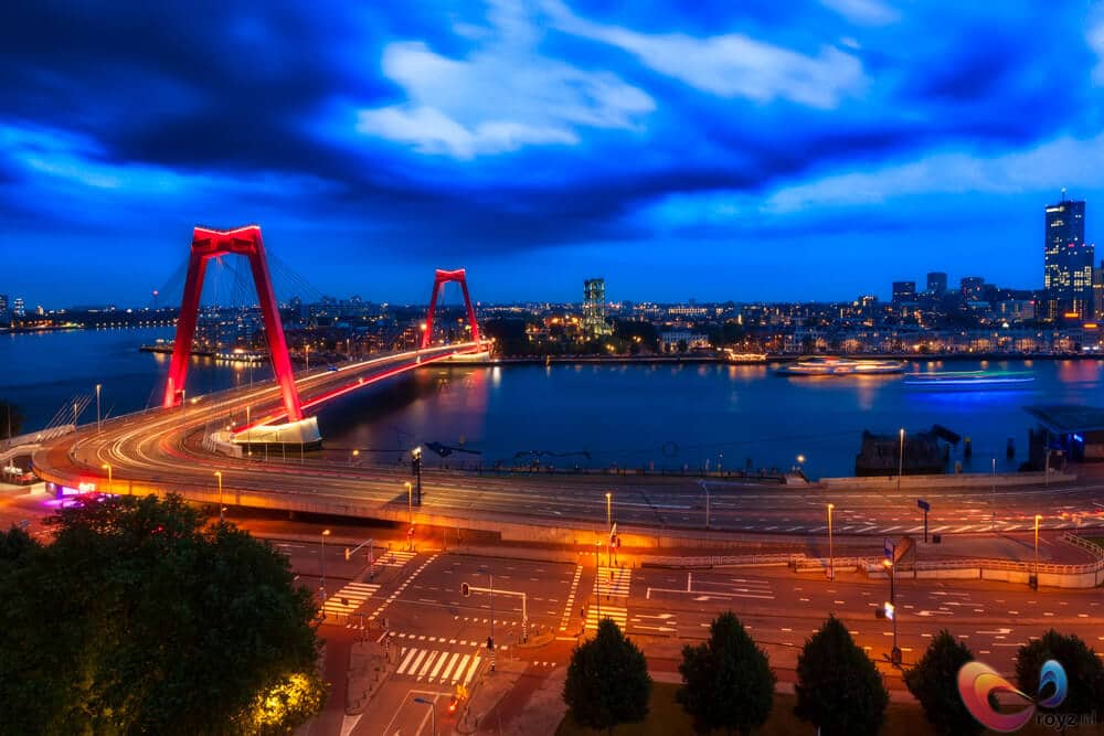 Rode brug bij blauw uur boven brede rivier met verlichte skyline.