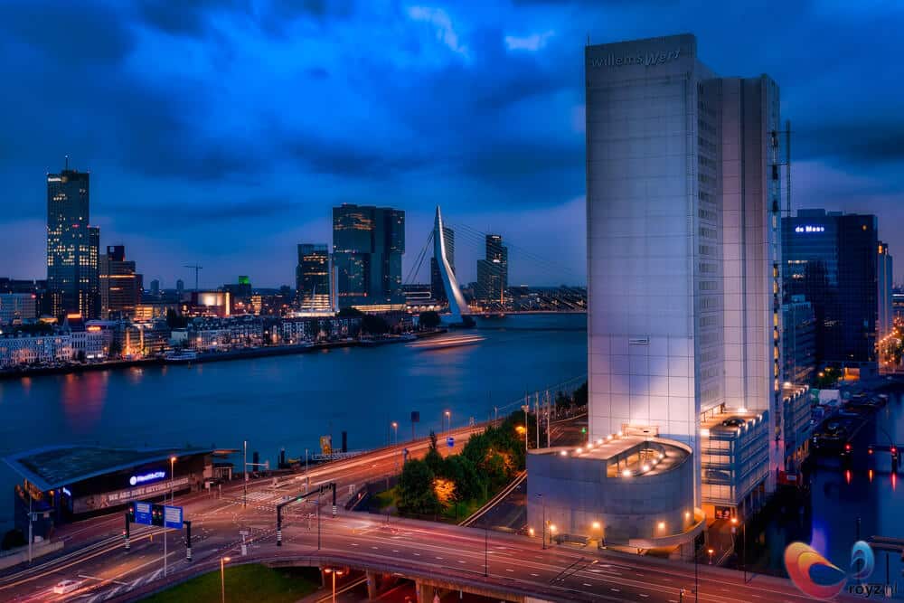 Rotterdam blauwe uur: Erasmusbrug, skyline en waterreflecties met warme ramen en lichtsporen.