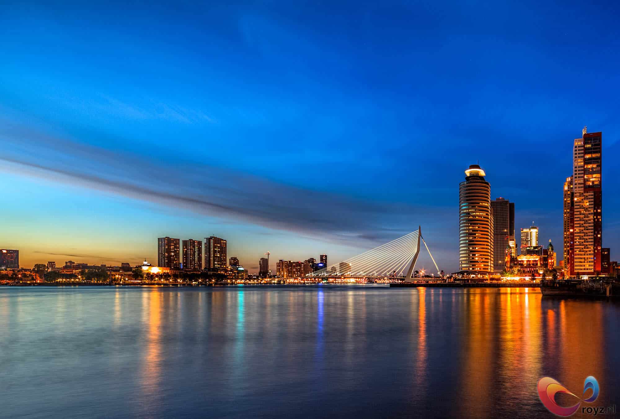 Erasmusbrug bij zonsondergang: Rotterdamse skyline met reflecties op een rustige rivier.