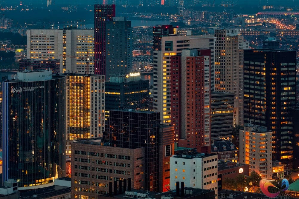 Rotterdamse skyline bij schemering: glanzende kantoortorens, havenlichten en nachtelijke bedrijvigheid.