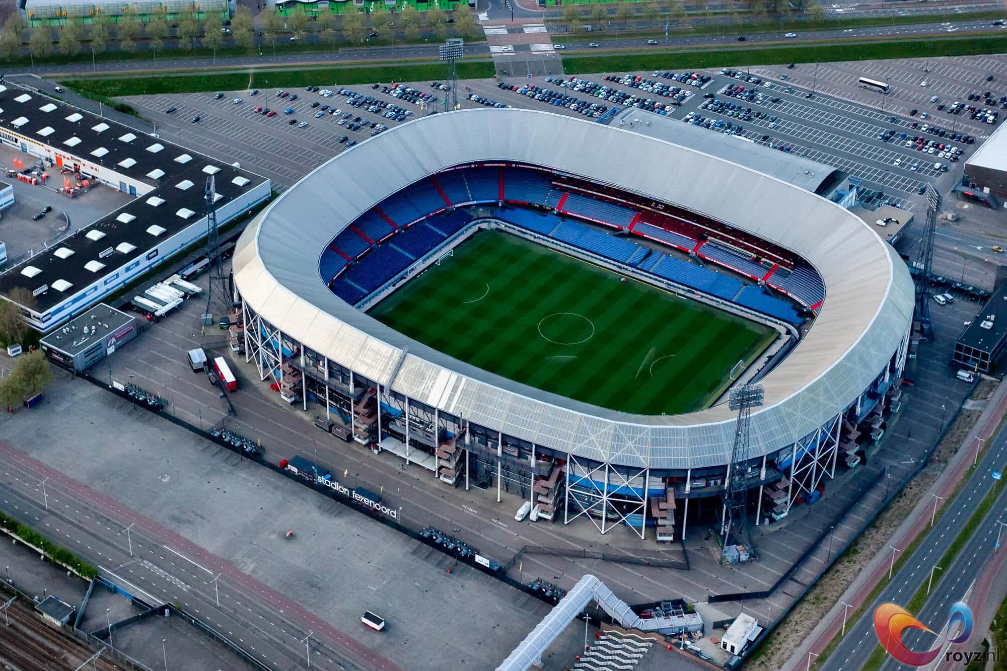 Luchtfoto van Rotterdam-stadion met blauwe en rode zitplaatsen, omgeven door parkeervoorzieningen en toegangswegen.